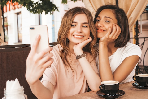 Two young beautiful smiling hipster girls in trendy summer clothes.Carefree women chatting in veranda cafe and drinking coffee.Positive models having fun and and taking selfie on smartphone