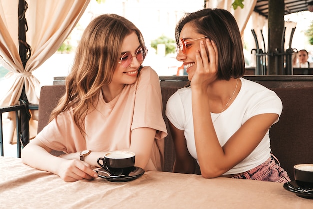 Two young beautiful smiling hipster girls in trendy summer clothes.Carefree women chatting in veranda terrace cafe and drinking coffee.Positive models having fun and communicating