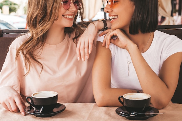 Due giovani belle ragazze sorridenti dei pantaloni a vita bassa in vestiti estivi d'avanguardia donne libere che chiacchierano nel caffè della terrazza della veranda e che bevono caffè modelli modulari divertendosi e comunicando