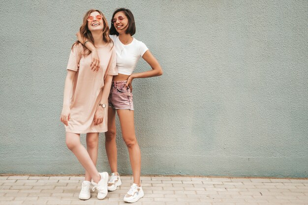 Two young beautiful smiling hipster girls in trendy summer clothes.Sexy carefree women posing near wall in the street in sunglasses. Positive models having fun and hugging