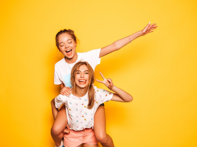 Two young beautiful smiling hipster girls in trendy summer clothes. Sexy carefree women posing near yellow wall.Model sitting on her friend's back and raising hands