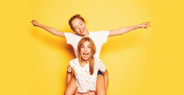 トレンディな夏服で2人の若い美しい笑顔流行に敏感な女の子。黄色の壁の近くでポーズセクシーな屈託のない女性。彼女の友人の背中に座って、手を上げるモデル