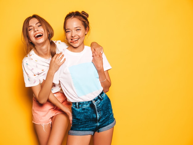 Two young beautiful smiling hipster girls in trendy summer clothes. Sexy carefree women posing near yellow wall. Positive models going crazy and having fun.Hugging