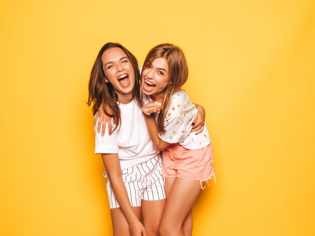 Two young beautiful smiling hipster girls in trendy summer clothes. Sexy carefree women posing near yellow wall. Positive models going crazy and having fun.Hugging