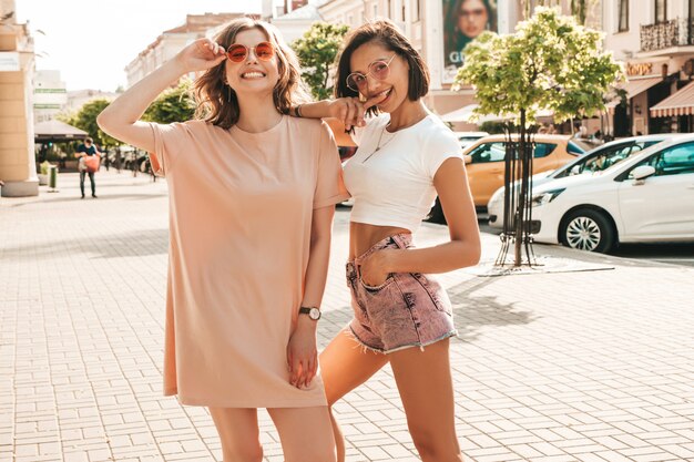 Две молодые красивые улыбающиеся хипстерские девочки в модной летней одежде. Сексуальные беззаботные женщины, позирующие на уличном фоне в солнцезащитных очках. Позитивные модели развлекаются и сходят с ума