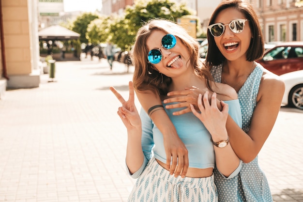 Две молодые красивые улыбающиеся хипстерские девочки в модной летней одежде. Сексуальные беззаботные женщины, позирующие на уличном фоне в солнцезащитных очках. Позитивные модели веселиться и обниматься. Показывать знак мира