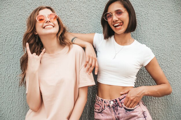 Две молодые красивые улыбающиеся хипстерские девочки в модной летней одежде. Сексуальные беззаботные женщины, позирующие на уличном фоне в солнцезащитных очках. Позитивные модели развлекаются и обнимаются