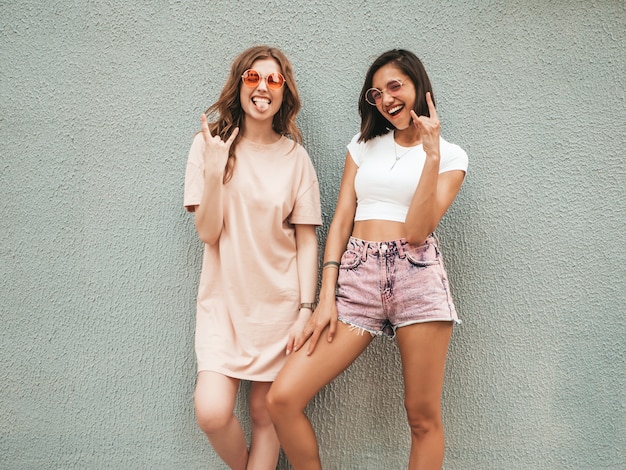 Две молодые красивые улыбающиеся хипстерские девочки в модной летней одежде. Сексуальные беззаботные женщины, позирующие на улице около стены в солнцезащитных очках. Позитивные модели развлекаются и показывают рок-н-ролл