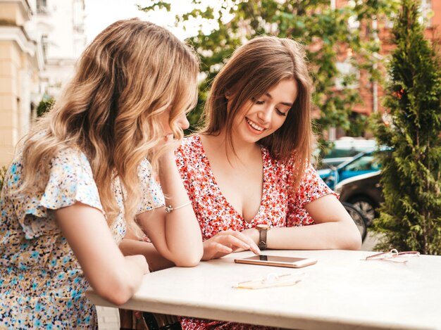 Две молодые красивые улыбающиеся хипстерские девушки в модном летнем сарафане. Беззаботные женщины, болтающие в кафе на веранде. Модель показывает информацию о своем друге в своем смартфоне. Они смотрят фотографии после покупок