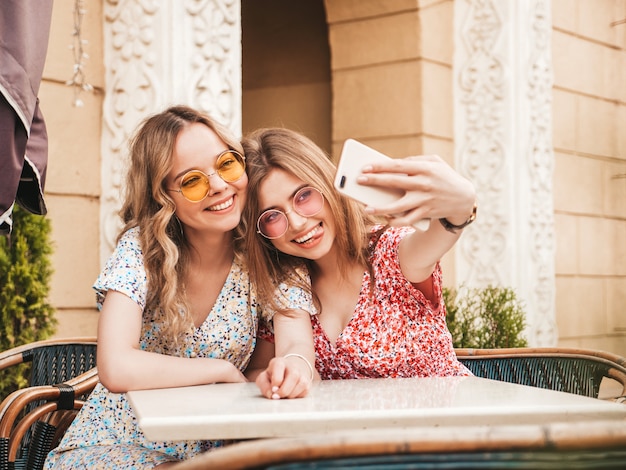 Two young beautiful smiling hipster girls in trendy summer sundress.Carefree women chatting in veranda cafe on the street background.Positive models having fun and and taking selfie on smartphone