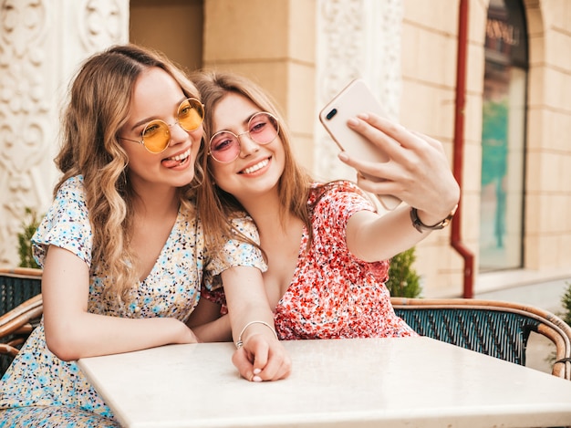 Two young beautiful smiling hipster girls in trendy summer sundress.Carefree women chatting in veranda cafe on the street background.Positive models having fun and and taking selfie on smartphone