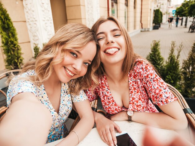 トレンディな夏のサンドレスで2人の若い美しい笑顔の流行に敏感な女の子。通りの背景にあるベランダカフェでチャットの屈託のない女性。