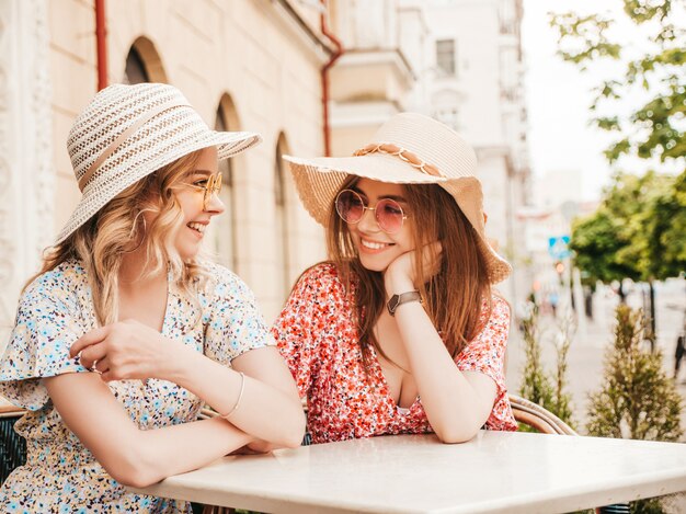 트렌디 한 여름 sundress에 두 젊은 아름 다운 미소 hipster 소녀 선글라스에서 거리 배경에 베란다 카페에서 채팅 평온한 여자. 재미 있고 의사 소통하는 긍정적 인 모델