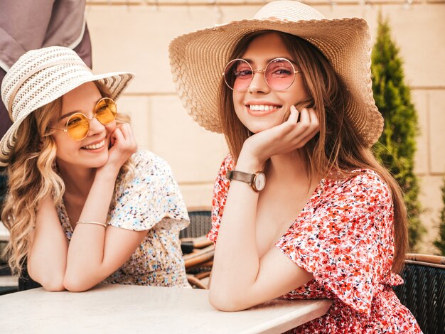 Two young beautiful smiling hipster girls in trendy summer sundress.Carefree women chatting in veranda cafe on the street background in sunglasses.Positive models having fun and communicating