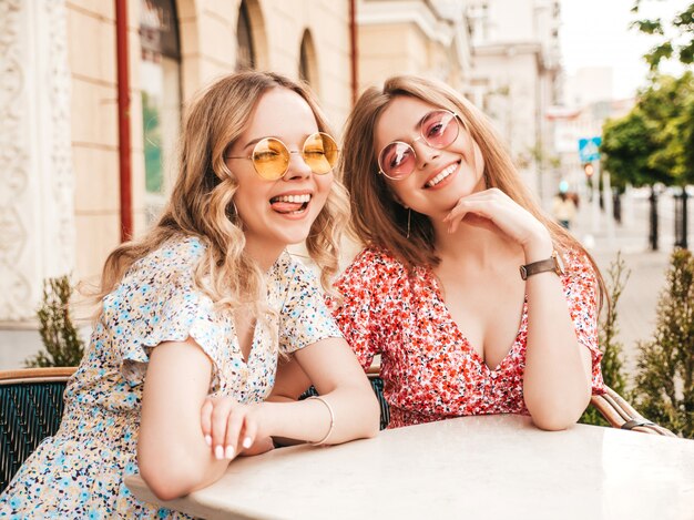 Two young beautiful smiling hipster girls in trendy summer sundress.Carefree women chatting in veranda cafe on the street background in sunglasses.Positive models having fun and communicating