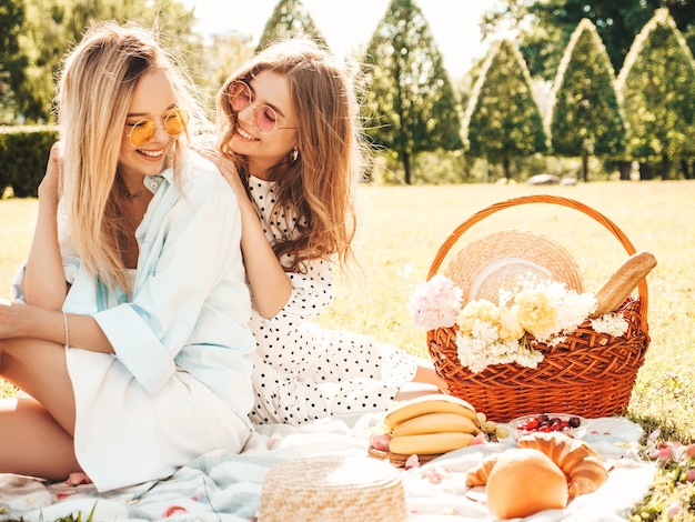 Две молодые красивые улыбающиеся хипстерские девушки в модном летнем сарафане и шляпах. Беззаботные женщины делают пикник на улице.