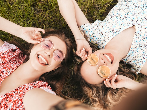 Two young beautiful smiling hipster girls in trendy summer sundress.Sexy carefree women lying on the green grass in sunglasses.Positive models having fun.Top view.Taking selfie photos on smartphone