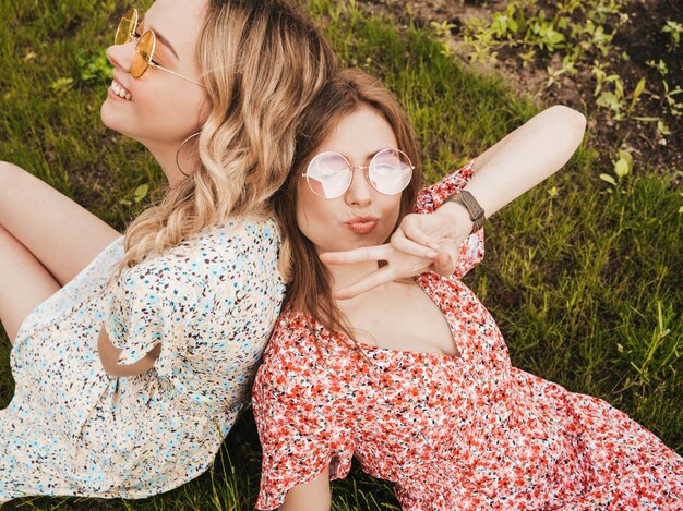 유행 여름 sundress에 두 젊은 아름 다운 웃는 hipster 여자. 선글라스에 푸른 잔디에 누워 섹시 평온한 여자. 재미있는 모델 재미.