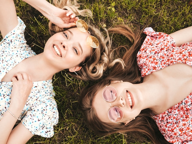 Two young beautiful smiling hipster girls in trendy summer sundress.Sexy carefree women lying on the green grass in sunglasses.Positive models having fun.Top view