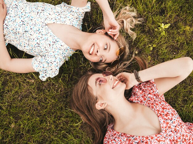 Две молодые красивые улыбающиеся хипстерские девушки в модном летнем сарафане. Сексуальные беззаботные женщины, лежащие на зеленой траве в солнцезащитных очках. Позитивные модели с удовольствием. Вид сверху
