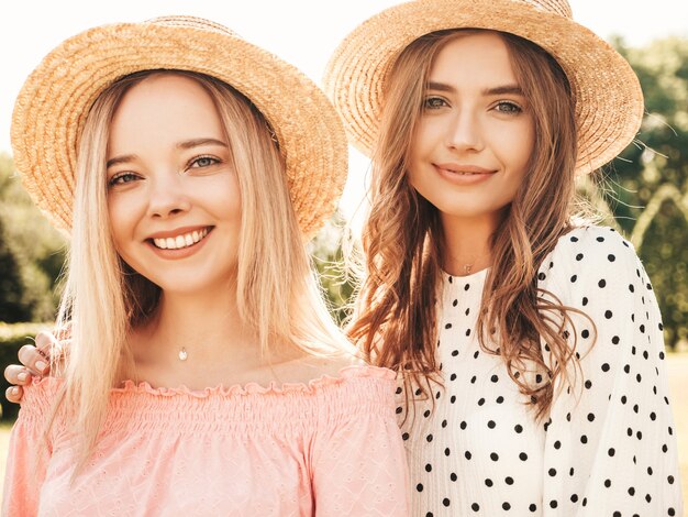 Two young beautiful smiling hipster girls in trendy summer sundress. Sexy carefree women posing in the park in hats. 