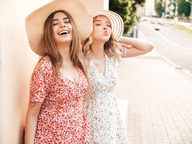 Two young beautiful smiling hipster girls in trendy summer sundress.Sexy carefree women posing on the street background in hats. Positive models having fun and hugging.They going crazy