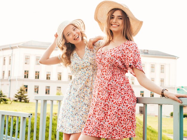 Two young beautiful smiling hipster girls in trendy summer sundress.Sexy carefree women posing on the street background in hats. Positive models having fun and hugging