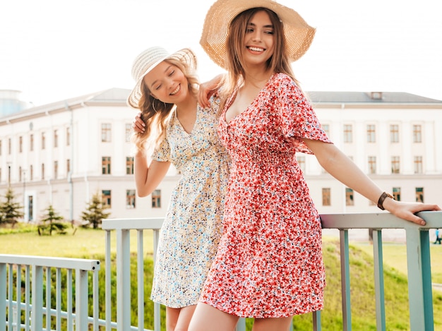 Две молодые красивые улыбающиеся битник девушки в модном летнем сарафане. Сексуальные беззаботные женщины, позирует на фоне улицы в шляпах. Позитивные модели развлекаются и обнимаются