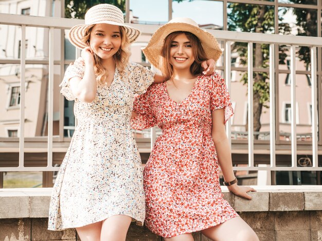 Two young beautiful smiling hipster girls in trendy summer sundress.Sexy carefree women posing on the street background in hats. Positive models having fun and hugging