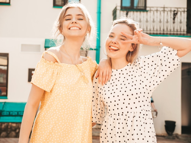 Две молодые красивые улыбающиеся битник девушки в модном летнем сарафане. Сексуальные беззаботные женщины, позирует на фоне улицы в солнцезащитные очки. Позитивные модели развлекаются и обнимаются