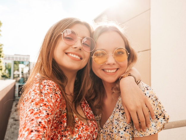 Two young beautiful smiling hipster girls in trendy summer sundress.Sexy carefree women posing on the street background in sunglasses. They taking selfie self portrait photos on smartphone at sunset