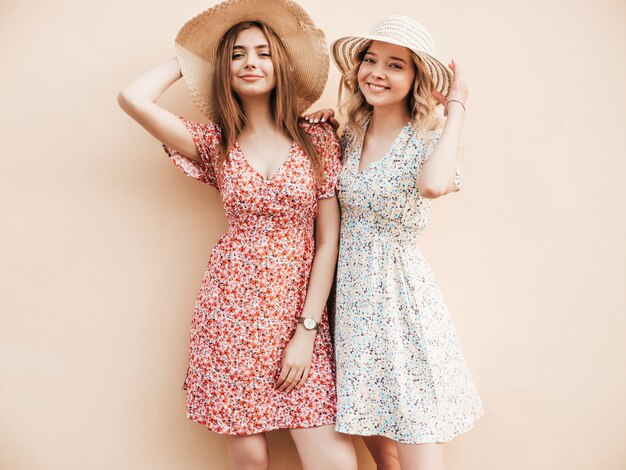 Two young beautiful smiling hipster girls in trendy summer sundress.Sexy carefree women posing on the street near wall in hats. Positive models having fun and hugging