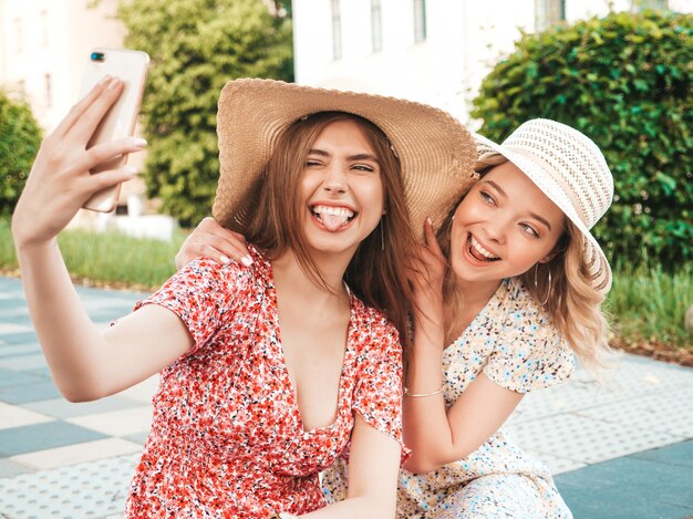 トレンディな夏のサンドレスで2人の若い美しい笑顔の流行に敏感な女の子。帽子で通りの背景に座っているセクシーな屈託のない女性。スマートフォンで自撮り写真を撮るポジティブモデル