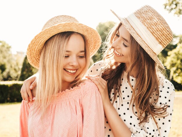 Two young beautiful smiling hipster woman in trendy summer sundress. Sexy carefree women posing in the park in hats. 