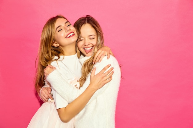 Two young beautiful smiling hipster women in trendy summer white clothes. Sexy carefree women posing near blue wall. Positive models hugging