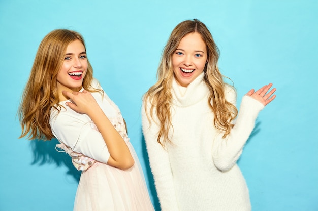 Two young beautiful smiling hipster women in trendy summer white clothes. Sexy carefree women posing near blue wall. Positive models