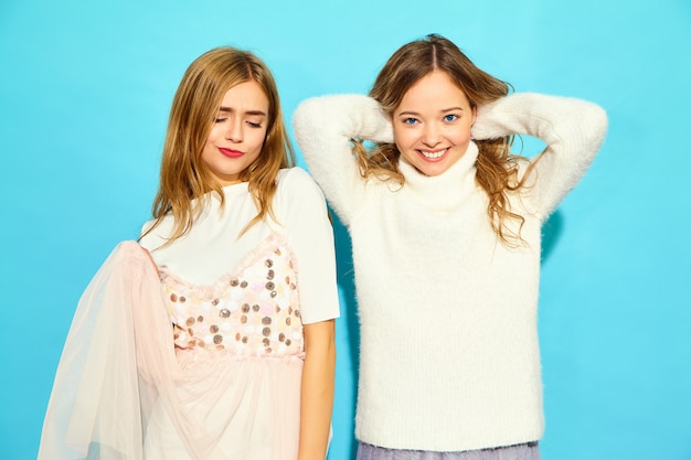 Two young beautiful smiling hipster women in trendy summer white clothes. Sexy carefree women posing near blue wall. Positive models