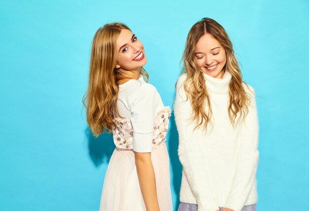トレンディな夏の白い服の2人の若い美しい笑顔流行に敏感な女性。セクシーな屈託のない女性が青い壁の近くでポーズします。ポジティブモデル