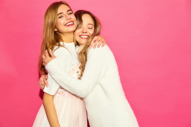 Two young beautiful smiling hipster women in trendy summer white clothes. Sexy carefree women posing near pink wall. Positive models hugging