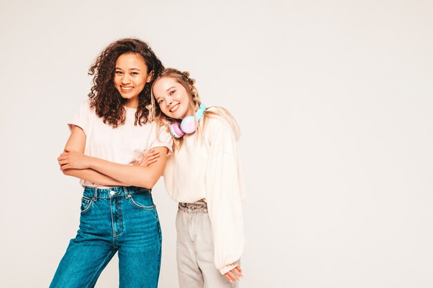 Two young beautiful smiling international hipster female in trendy summer clothes. carefree women posing on grey