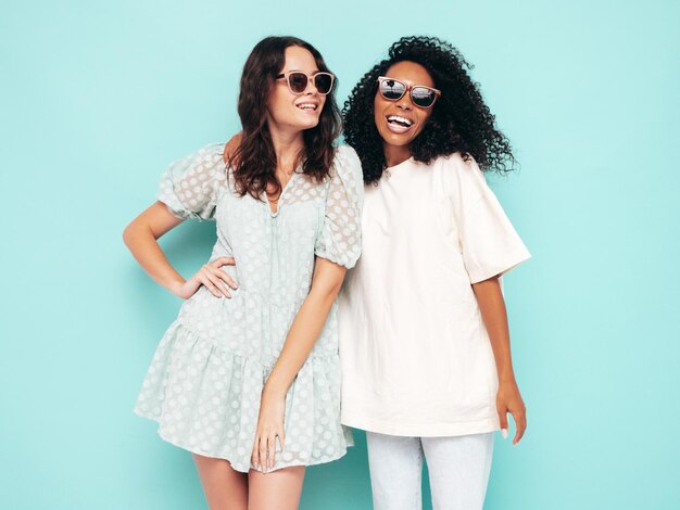 최신 유행의 여름 옷을 입은 두 명의 젊은 아름다운 국제 힙스터 여성 스튜디오의 파란색 벽 근처에서 포즈를 취하는 섹시하고 평온한 여성 우정의 개념을 즐기고 있는 긍정적인 모델