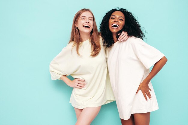 최신 유행의 여름 옷을 입은 두 명의 젊은 아름다운 국제 힙스터 여성 스튜디오의 파란색 벽 근처에서 포즈를 취하는 섹시하고 평온한 여성 우정의 개념을 즐기고 있는 긍정적인 모델