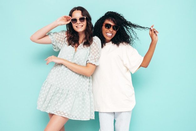 최신 유행하는 여름 옷을 입고 웃고 있는 두 젊은 국제 힙스터 여성 섹시하고 평온한 여성 스튜디오의 파란색 벽 근처에서 포즈를 취하는 긍정적인 모델