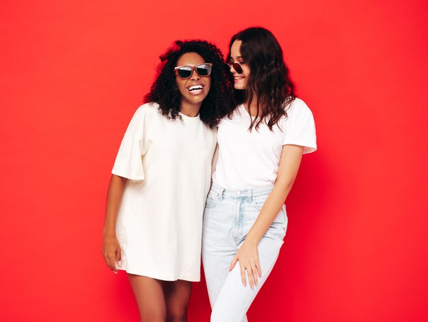 최신 유행의 여름 옷을 입고 웃고 있는 두 젊은 국제 힙스터 여성 섹시하고 평온한 여성 스튜디오의 빨간 벽 근처에서 포즈를 취하는 긍정적인 모델 선글라스에서 우정의 개념을 재미있게 즐기고 있습니다.
