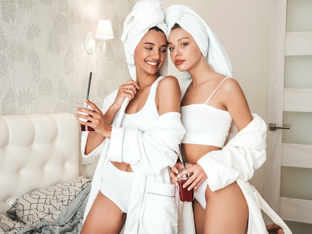 흰 목욕 가운과 머리에 수건을 입은 두 젊은 아름다운 웃는 여성. 호화로운 아파트나 호텔 방에서 침대에 앉아 있는 섹시하고 평온한 모델. 그들은 신선한 칵테일 스무디를 마시고 있다