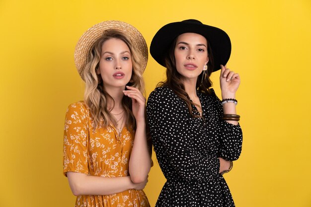 Two young beautiful women friends together isolated on yellow background in black and yellow dress and hat stylish boho trend spring summer fashion style accessories confident sexy