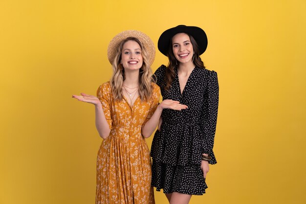 Two young beautiful women friends together isolated on yellow background in black and yellow dress and hat stylish boho trend, spring summer fashion style accessories, smiling happy mood having fun