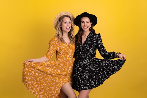 Two young beautiful women friends together isolated on yellow background in black and yellow dress and hat stylish boho trend spring summer fashion style accessories smiling happy mood having fun