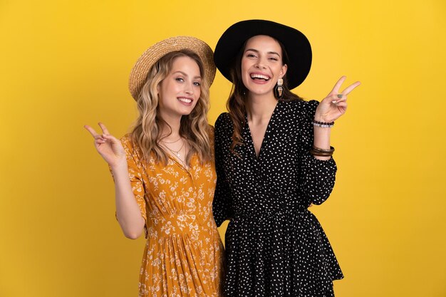 Two young beautiful women friends together isolated on yellow background in black and yellow dress and hat stylish boho trend spring summer fashion style accessories smiling happy mood having fun