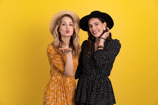 Two young beautiful women friends together isolated on yellow background in black and yellow dress and hat